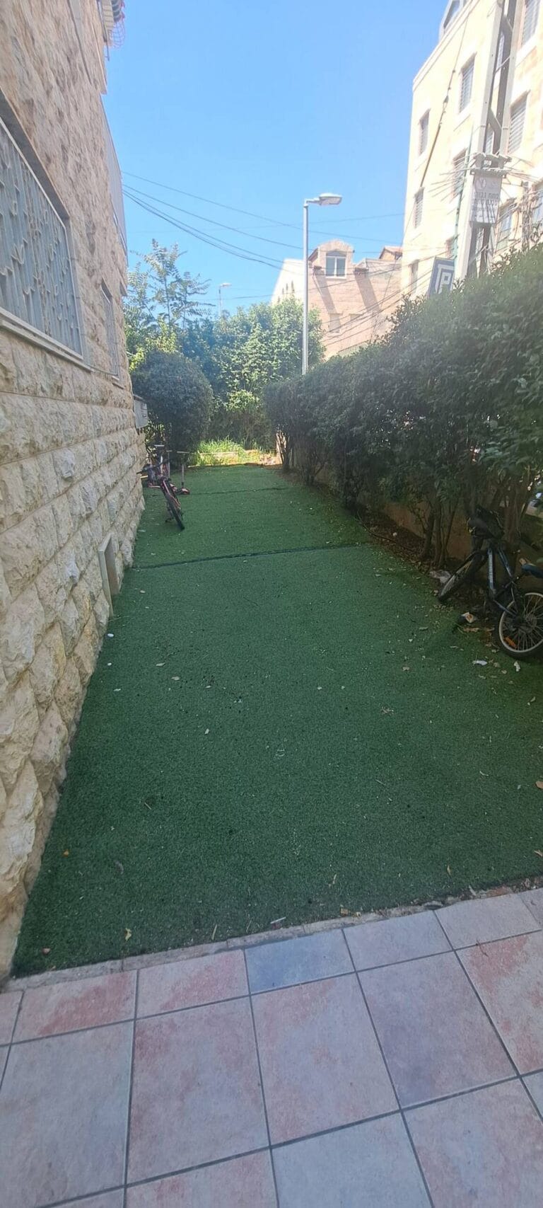Narrow outdoor garden with green ground covering beside a stone wall at the Hakalir 4-bedroom Rechavia apartment.
