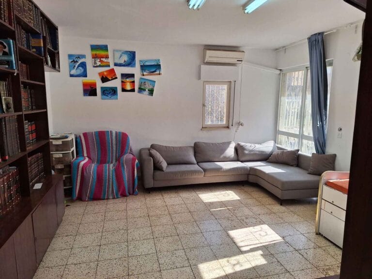 Living room with a gray corner sofa, striped armchair and bookshelves in the Hakalir 4-bedroom apartment, Rechavia, Jerusalem.