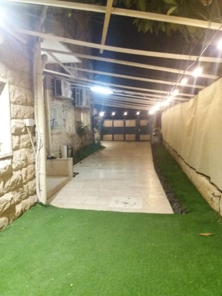 Paved courtyard with artificial grass, overhead framework and evening string lights at Ramban Giant Sukkah Apartment, Jerusalem
