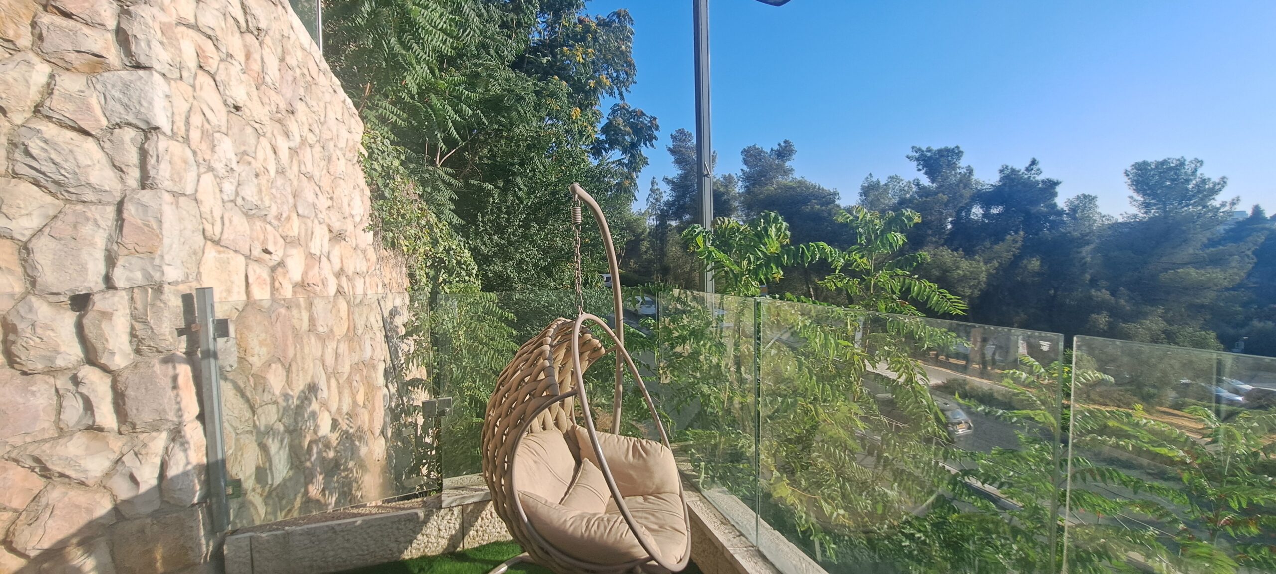 Hanging garden chair beside the glass railing and green Gan Sacher outlook at Wolfson Villa in Jerusalem.