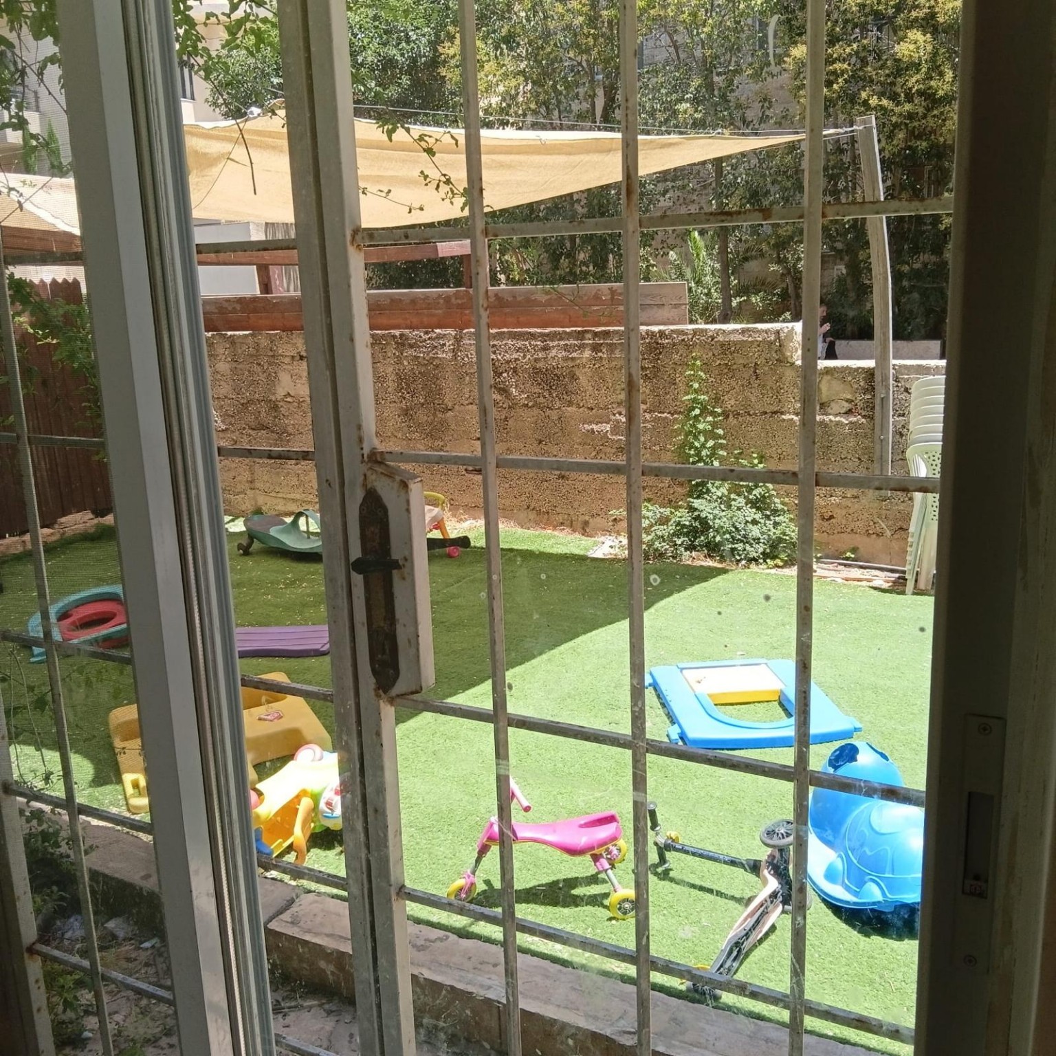 View through a metal grille onto artificial grass, greenery and children's toys at Ibn Shaprut Apartment #2