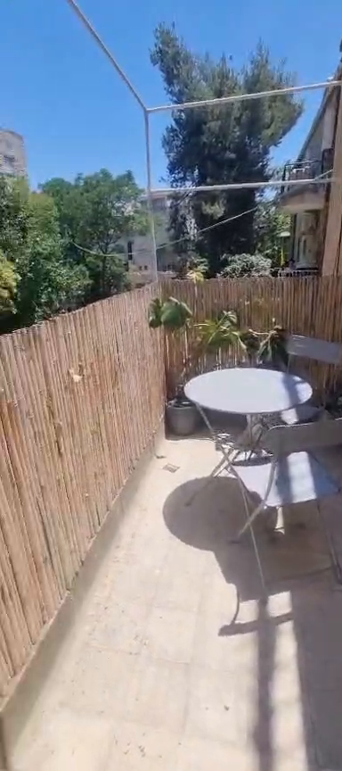 Balcony with a round table, folding chairs, bamboo screening and greenery at the Ibn Shaprut apartment.