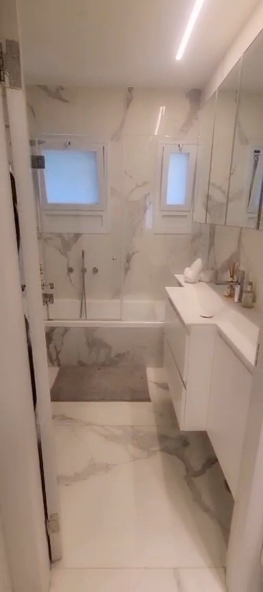 Bathroom with a white vanity, mirror, bathtub and gray-veined wall tiles in the Ibn Shaprut apartment.