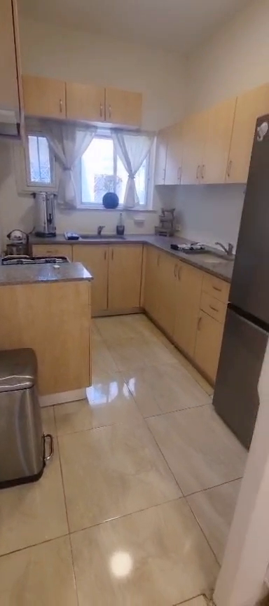 Kitchen with wood-tone cabinets, tiled floor, window and refrigerator in the Ibn Shaprut apartment.