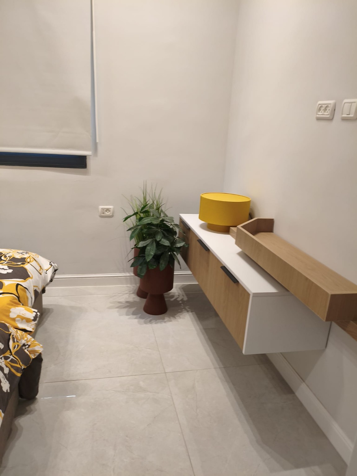 Floating wood-and-white bedroom console with a yellow lamp and potted plant in the Ramban Rechavia apartment.