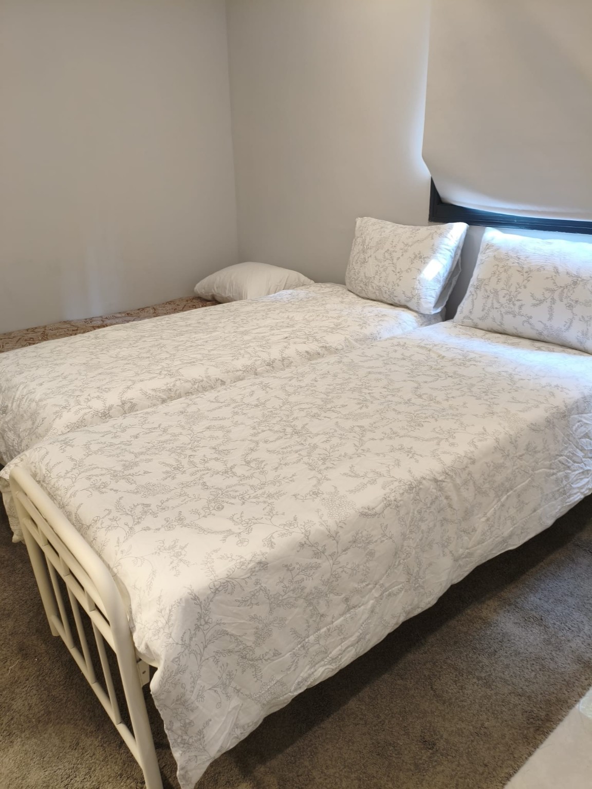Beds with white floral bedding and white metal frames beside a window in the Ramban Rechavia apartment.