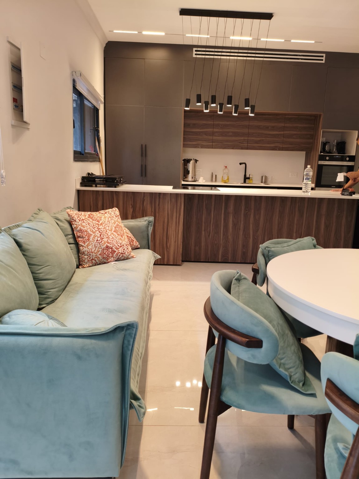 Teal sofa and dining chairs beside a round table, facing the wood-fronted kitchen island in the Ramban Rechavia apartment.