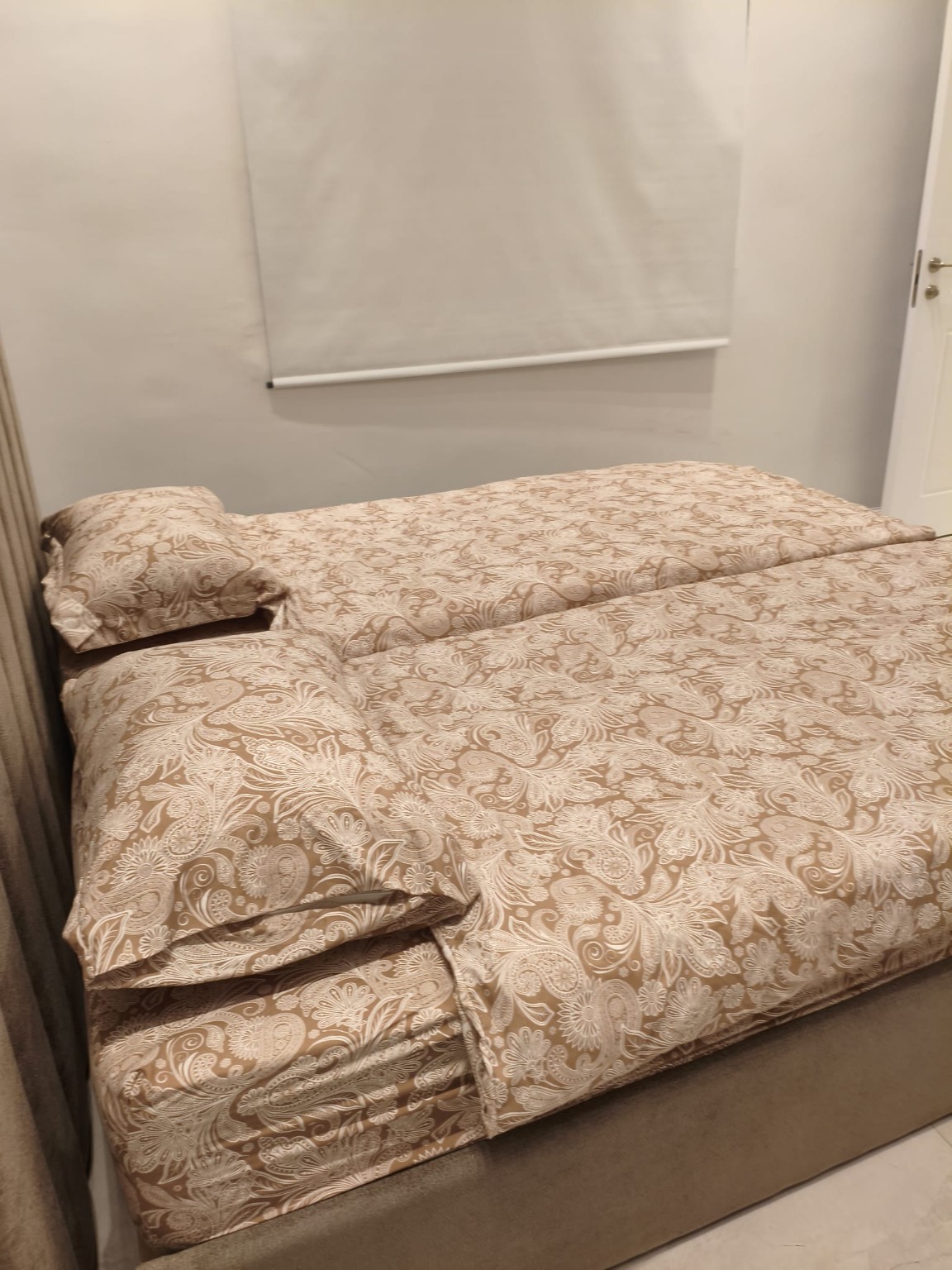 Two separate beds with matching taupe paisley bedding beneath a roller blind in the Ramban Rechavia apartment.